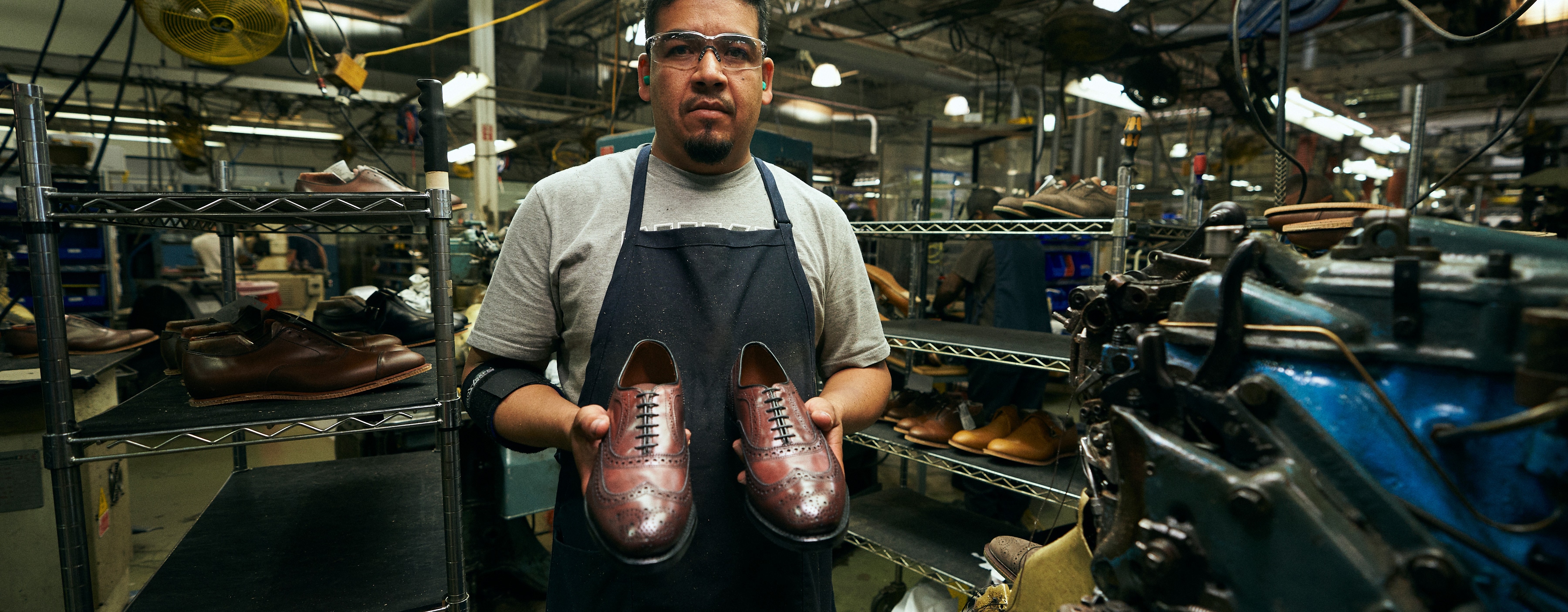 Image of a man in a shoe factory wearing safety goggles and ear plugs and holding up a pair of brown dress shoes